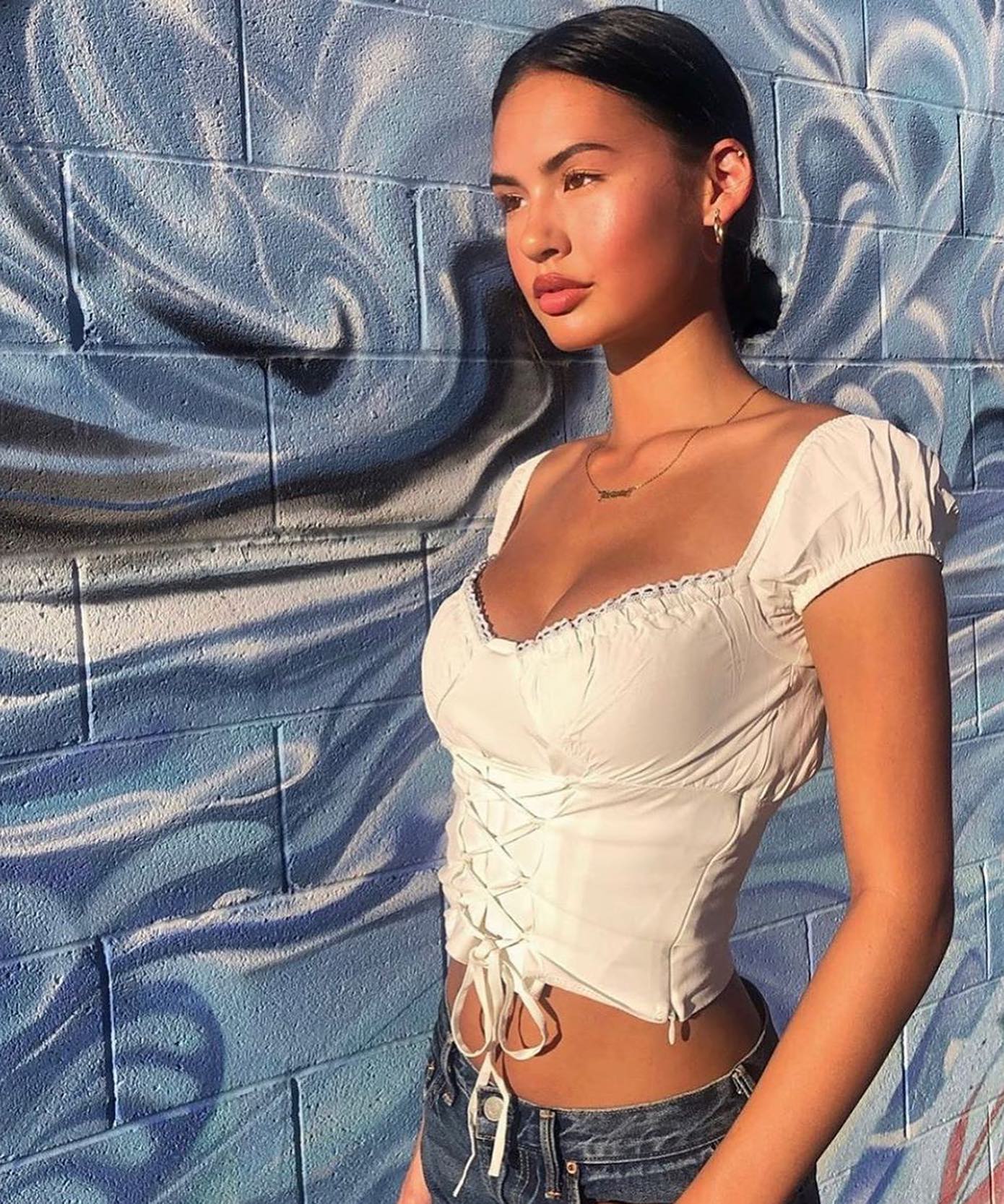 Woman in a white top and denim shorts standing against a textured blue wall.