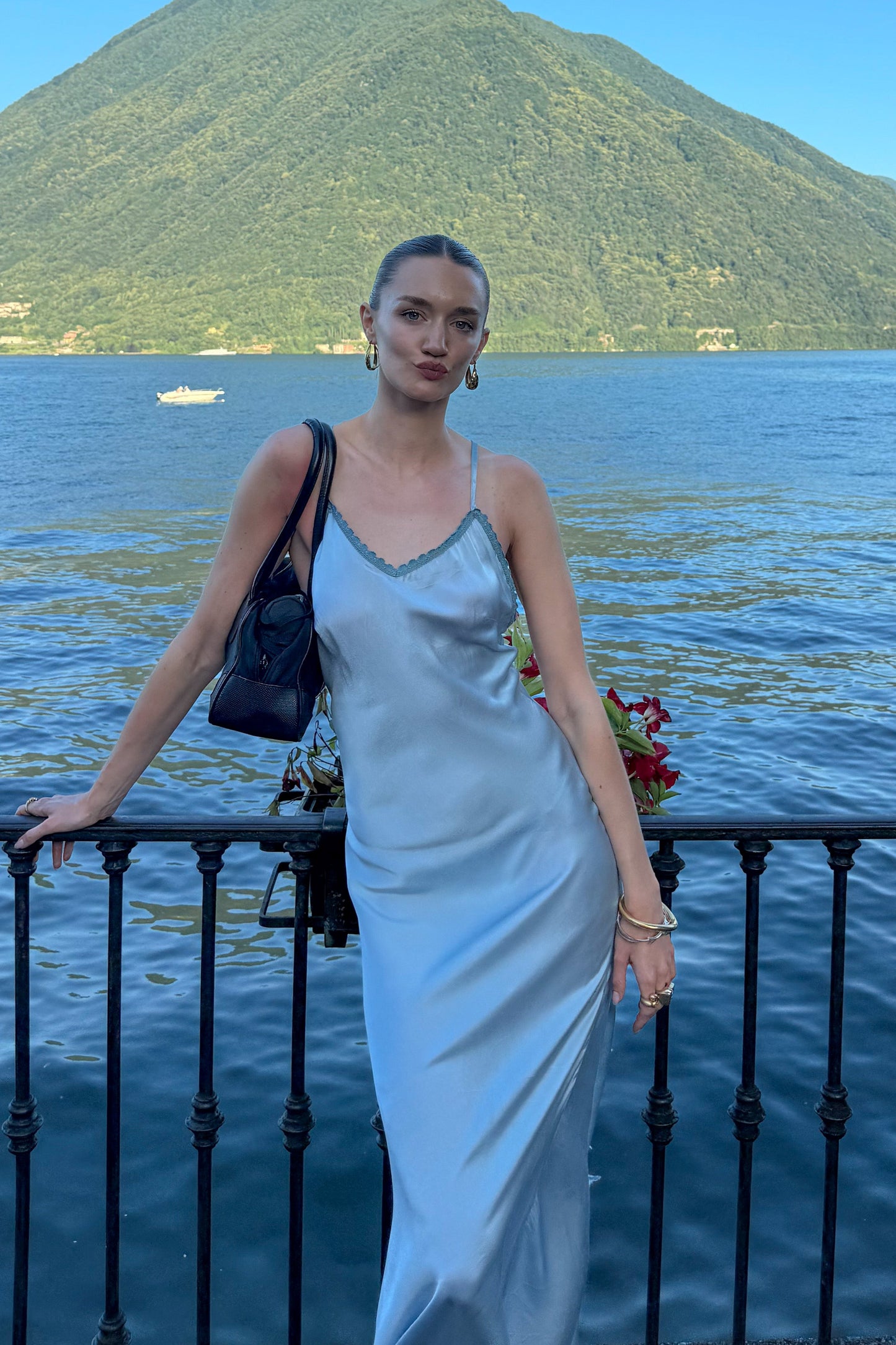 Woman in a light blue dress standing by a lake with a mountain in the background.