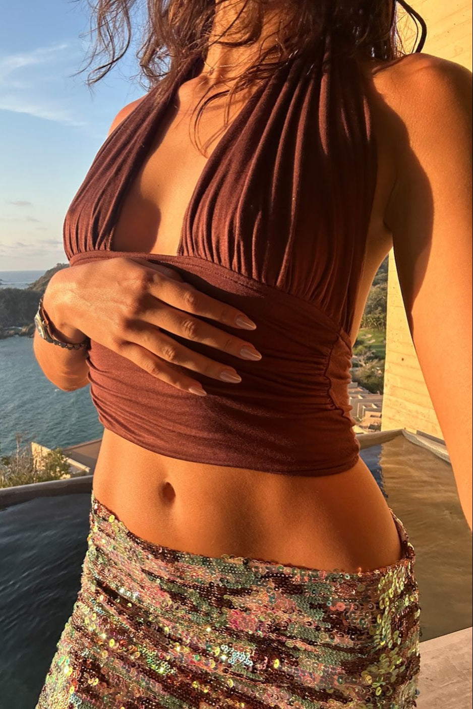 Woman in a brown halter top and patterned skirt standing by a pool with a scenic view.