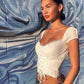 Woman in a white top and denim shorts standing against a textured blue wall.