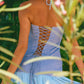 Woman in a blue dress with a lattice back standing among green foliage and pink flowers.
