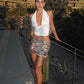 Woman in a white top and patterned skirt standing on a pathway with buildings and palm trees in the background.